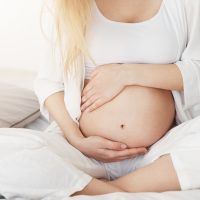 Young pregnant lady touching her belly sitting in her bedroom in afternoon. Pregnancy concept.
