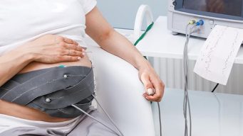 Pregnant woman performing cardiotocography (CTG). The belt on her belly connected to the Cardiotocograph machine aka Electronic Fetal Monitor (EFM) recording fetal heartbeat and uterine contractions